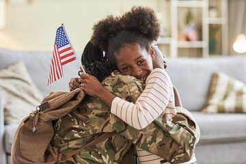 Portrait of African American little daughter with american flag embracing her mom in military uniform and smiling at camera