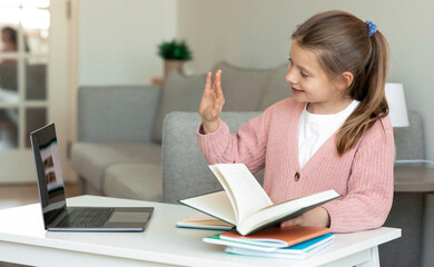 Smiling european teenager girl doing homework and reading book, waving hand at camera computer