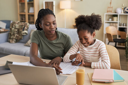 African American mother helping her daughter with homework while they sitting at table with laptop in the living room