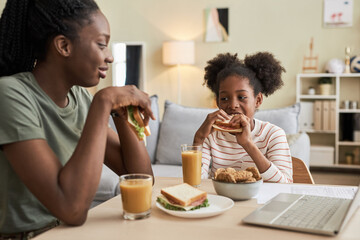 Mother eating sandwich with juice together with her little daughter at table in the room