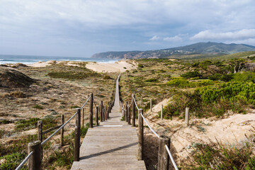 Fototapeta na wymiar Empty, wooden boardwalk on a beach Praia do Guincho in Sintra. View of grass and sand with hills and Atlantic Ocean in the background