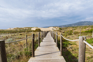 Empty, wooden boardwalk on a beach Praia do Guincho in Sintra. View of grass and sand with hills and Atlantic Ocean in the background - obrazy, fototapety, plakaty