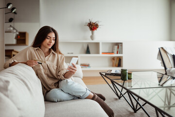 Obraz premium Positive millennial european lady shopaholic with credit card looking at phone, order purchases