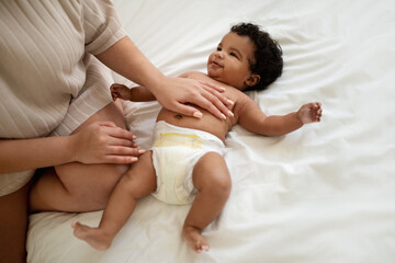 Millennial african american woman stroking belly of little baby on white bed in bedroom, top view