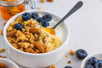 Bowl of oat granola with yogurt, banana, blueberries, chia seeds, honey and nuts on white background