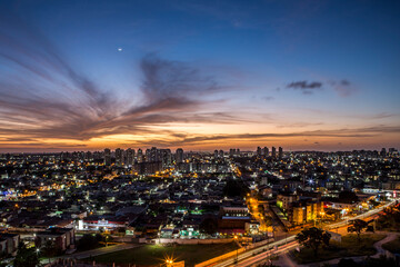 Fototapeta premium Sunset seen from above the building with orange and blue colored sky with the moon in the center and avenues and buildings highlighted. | Avenida Ayrton Senna em Natal, RN.