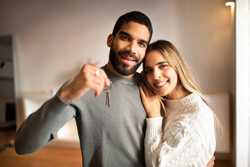 Positive young multiethnic husband and wife show keys from new apartment, enjoy buy own home in room