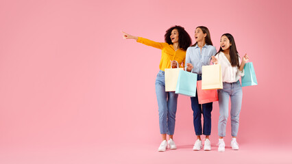 Three excited diverse ladies with shopping bags pointing and looking aside, pink background,...