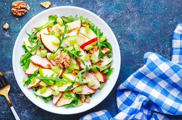Waldorf salad with green and red apple, celery, lettuce, chicken fillet, arugula and crunchy walnuts on plate, blue table background, top view