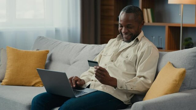 Mature smiling african american man paying online with credit card using laptop on couch at home happy male customer shopper pay for purchase on internet using e-banking service computer application