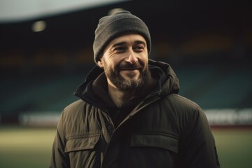 Portrait of a smiling bearded man in a warm jacket and hat standing in the stadium.