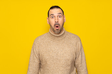 Bearded hispanic man in his 40s wearing a beige turtleneck tremendously amazed and surprised with open mouth in shock, isolated over yellow background.