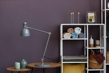 Lamp and two small vases standing on round wooden tables by wall against rack of shelves with rolled towels, picture frames and basket