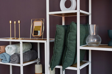 Part of shelf rack with two dark green cushions, picture frames and holders, rolled soft towels, vases and other stuff standing by wall of domestic room
