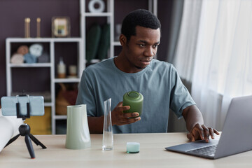 Young male blogger holding handmade vase while sitting by workplace in front of laptop and looking at screen during online masterclass