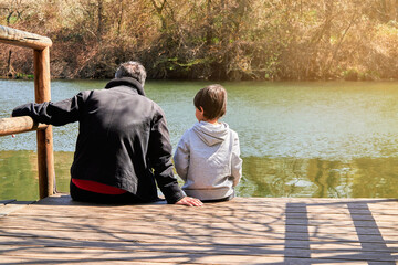 Unrecognizable father and son stays sitting on a dock over a river