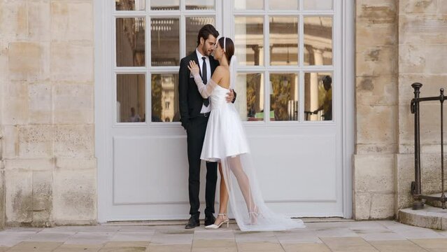 Happy newlyweds embracing each other by the white entrance door with windows. Action. Young and pretty man and woman in suit and white short dress.