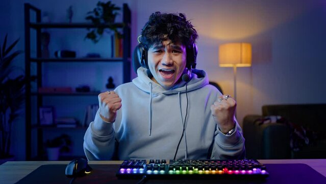 Young Asian man in headphones plays computer game, using computer mouse and keyboard, winning match