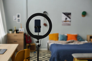Focus on round frame with smartphone standing in the center of bedroom with double bed with several cushions, chair with backpack and desk