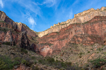 Scenic view on a massiv cliff seen from Bright Angel hiking trail at South Rim of Grand Canyon National Park, Arizona, USA. Light shining on steep stone wall and rock formations with blue sky