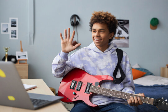 Happy teenager in casualwear greeting his music teacher by waving hand while sitting on bed in front of laptop before online lesson