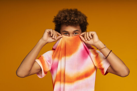 Cute teenage boy hiding face behind colorful t-shirt while standing in front of camera against yellow background during photo session