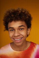 Face of cute smiling teenage boy in trendy t-shirt looking at you while standing in front of camera against yellow background in studio