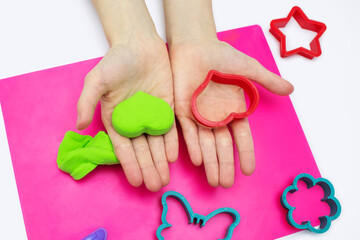 The child plays with soft green plasticine and a heart-shaped mold. Hands of a child with airy plasticine and close-up in the shape of a heart. A set of different molds for modeling plasticine