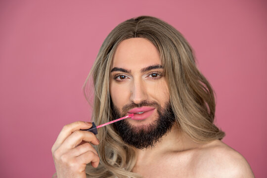 Transgender wearing blonde wig and white dress doing makeup applying lip gloss preparing for party