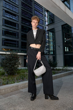 Full body shot of red head gay man in black suit and cowboy boots holding a silver sparkly handbag walking through streets of city near business buildings and posing for photo with confidence outdoors