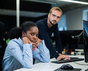 Colleagues look at the monitor and decide working moments. Caucasian man helps sad african woman solve computer problem.