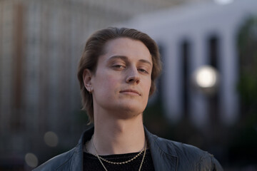 Expressing tranquility, happiness, and their queer, fluid gender identity, a genderfluid model poses with a calm, direct gaze while standing on the sidewalk in the later afternoon sun.