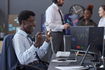 African American broker happy with successful deal during conversation on the phone sitting at his workplace with computers