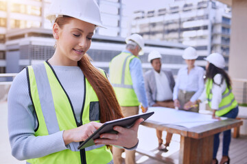 Construction is underway. an attractive young female construction worker using a tablet while standing outside with her team in the background.