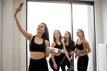 Fototapeta na wymiar Attractive healthy women with sports bottles taking selfie on cell phone while resting after yoga class in fitness centre. Cheerful friends in modern activewear enjoying healthy lifestyle together.