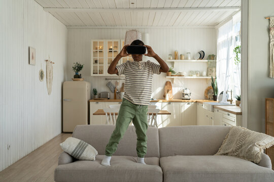 Interested african american boy plays video game stands on couch at home in virtual reality helmet. Interested preteen watches movie in vr goggles. Virtual reality, cyberspace, entertainment concept.