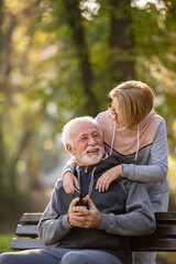 Active modern cheerful senior couple meet in the park. An older man is sitting on a park bench using smartphone, and an elderly woman surprises him from the back.