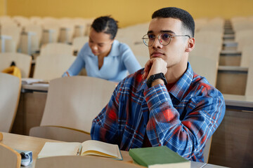 Teenage boy in eyeglasses sitting at desk and listening to lecturer during lecture in university