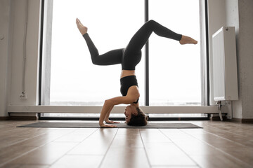 Relaxed dark haired woman using yoga mat for doing Pincha Mayurasana exercise at spacious fitness studio. Caucasian female in sport attire training regularly for body care.