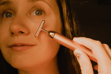 Obraz premium Young beautiful white woman, girl is using vibrating golden facial massager on dark black background with copy space. Preservation prolongation of natural beauty youth Female skin care with pleasure.