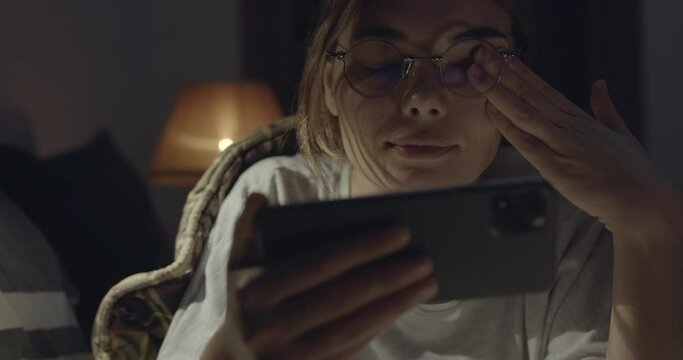 Tired woman using mobile phone and rubbing her eyes while laying on bed at night