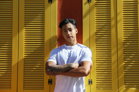 Handsome young latin man in plain white t-shirt standing near the rustic house with red walls and yellow window shutters. Close up, copy space, background.