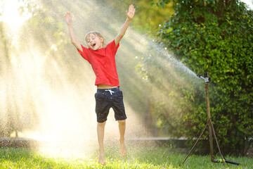 Funny little boy playing with garden sprinkler in sunny city park. Elementary school child laughing, jumping and having fun with spray of water. Summer outdoors activity for kids. - obrazy, fototapety, plakaty
