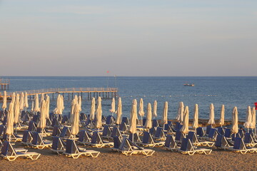 Beach with sun loungers and umbrellas in the rays of the setting sun on the turquoise Mediterranean Sea in Alanya, Turkey, April 2023.