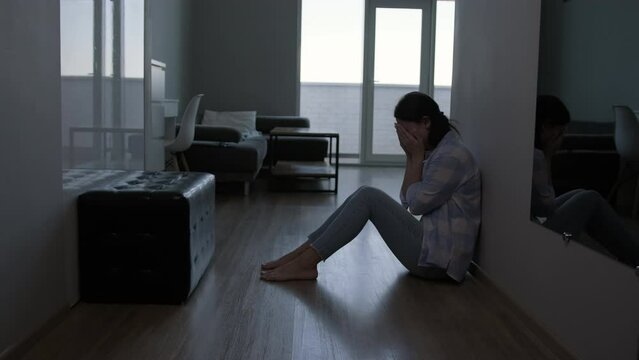 A woman cries after reading a message on a smartphone while sitting on the floor at home.