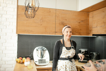 Portrait of young smiling beauteous woman wearing dirty white apron with dots, grey sweatshirt, standing in kitchen.