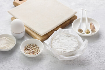 Ingredients for a delicious homemade pie or snack: puff pastry, white mold cheese (camembert or brie), egg, spices, seed mixture and flour on a light gray background.