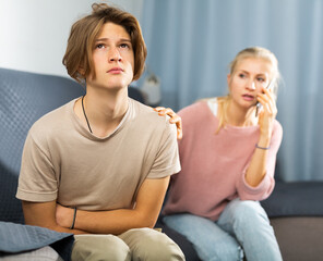 Teenager with stomach ache sitting on couch at home and holding his stomach with hands while worried mom calling doctor on phone