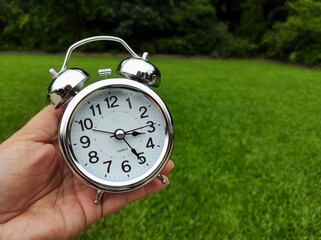 Antique silver alarm clock in human hand on blurred grass background.