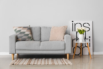 Cozy sofa with cushions and tulip flowers in vase on table near grey wall